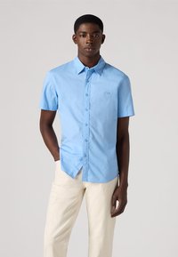 Lichtblauw kortoe-shirt met een button-down kraag, gestructureerde stof, en geborduurd logo op de linkerborst, gedragen met lichtgekleurde broek.