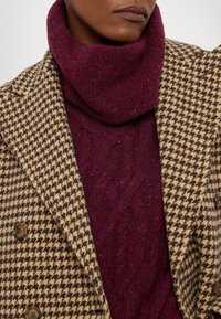 Plašč v houndstooth vzorcu v rjavi in bež barvi z dvema gumboma, položen čez strukturirano bordo pulover s turtleneck ovratnikom.