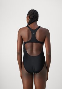 Crni jednodijelni kupaći kostim izrađen od glatkog materijala, s dizajnom s racerback-om i prozirnim stražnjim panelom za elegantan izgled. Puma logo na gornjem dijelu leđa.