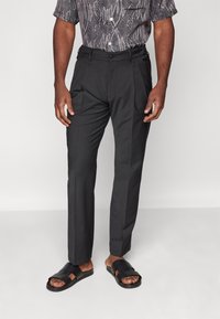 Pantalons noirs ajustés en tissu lisse, avec des plis et des poches latérales, associés à des sandales noires à glissière avec des brides texturées.