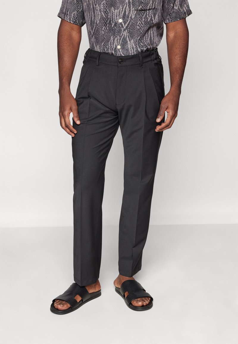 Pantalons noirs ajustés en tissu lisse, avec des plis et des poches latérales, associés à des sandales noires à glissière avec des brides texturées.