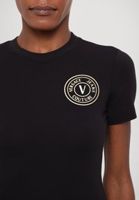 Juoda trumpa rankovė T-marškiniai, pagaminti iš medvilnės, su apvaliu auksiniu logotipu, ant kurio užrašyta "VERSACE JEANS COUTURE" ir centrinis "V" dizainas.