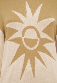 Μπεζ πλεκτό πουλόβερ με μεγάλο, εκρού σχέδιο ηλιακής ακτίνας, με κυκλικό κέντρο και μυτερές ακτίνες, που αναδεικνύει μαλακό,  υφή ύφασμα.