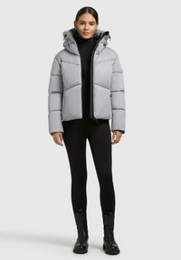 Una donna che indossa un giubbotto imbottito grigio chiaro con cappuccio, dolcevita nera, leggings neri e stivaletti neri mentre è in piedi contro uno sfondo bianco.