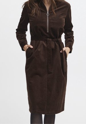 Femme portant une robe en velours côtelé marron foncé à manches longues, avec une fermeture éclair à l'avant, une ceinture à la taille, et les mains dans les poches, sur un fond uni.