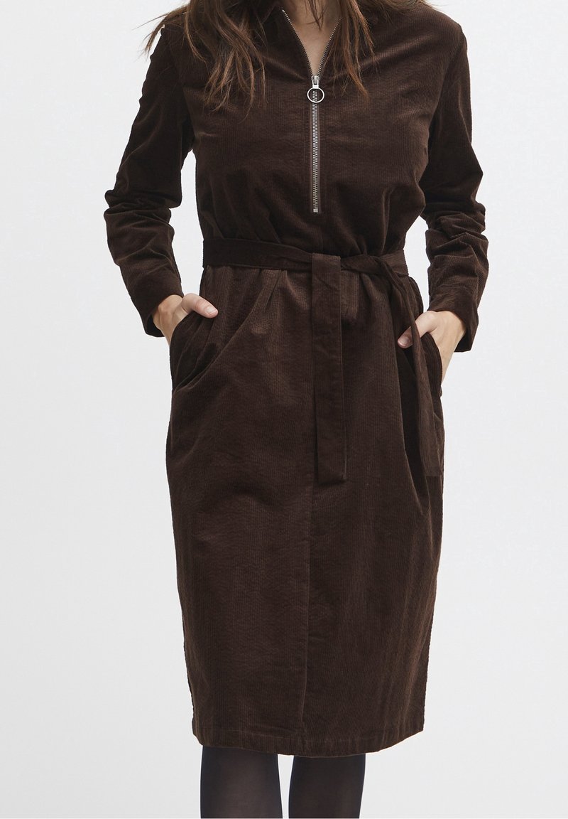Femme portant une robe en velours côtelé marron foncé à manches longues, avec une fermeture éclair à l'avant, une ceinture à la taille, et les mains dans les poches, sur un fond uni.