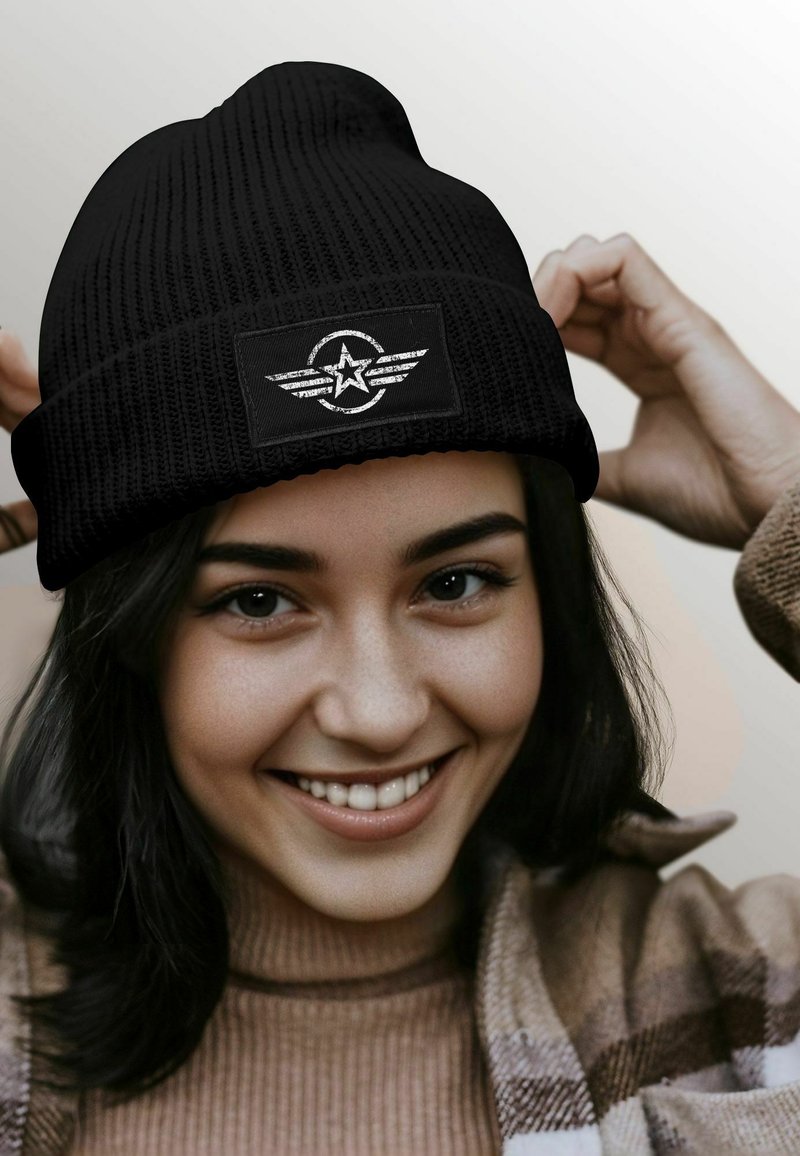 Black knit beanie featuring a textured fabric and a rectangular patch with a white star and wing design, worn by a smiling person.