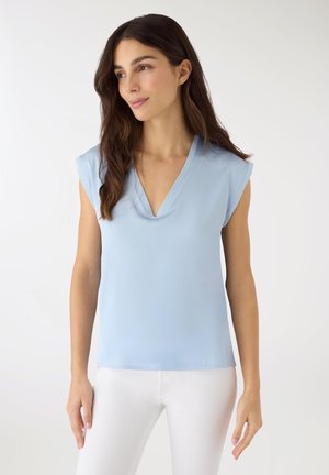 Donna con capelli lunghi e scuri che indossa una maglietta smanicata azzurro chiaro con scollo a V e pantaloni bianchi, che guarda leggermente di lato su uno sfondo semplice.