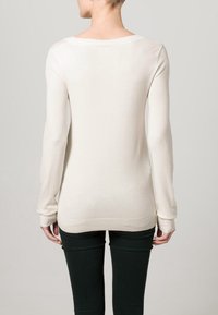 Maglione a maglia color crema con maniche lunghe e scollatura ampia, caratterizzato da una texture liscia e polsini a costine. Indossato con pantaloni aderenti verdi scuri.