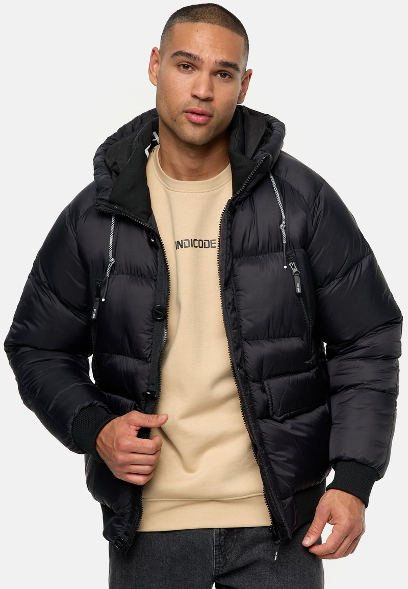 Sort pufferjakke med hætte, der har lynlås foran, to brystlommer og elastiske manchetter. Båret over en beige sweatshirts.