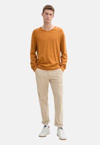 Suéter de punto naranja quemado de manga larga y cuello en V combinado con pantalones beige y zapatillas blancas. El suéter tiene un corte holgado y puños acanalados.