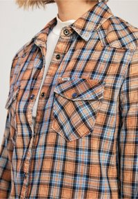 Camicia a quadri nei toni dell'arancione, blu e nero. Presenta due tasche sul petto, chiusura a bottoni e una consistenza di tessuto morbido.