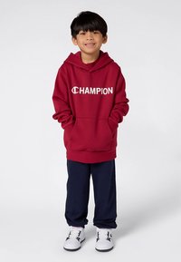 Sudadera burdeos con capucha y bolsillo delantero, con el logo blanco de "CHAMPION". Combinada con pantalones deportivos azul marino y zapatillas blancas.