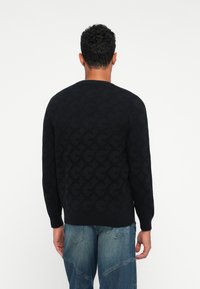 Mann in einem schwarzen Langarmpullover mit gemusterten "G"-Buchstaben und blauen Patchwork-Jeans, der mit dem Rücken zu einer weißen Hintergrund steht.