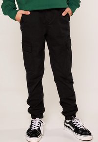 Černé cargo kalhoty s bočními kapsami, slim fit, vyrobené z bavlněné směsi; nošeny s černobílými teniskami s tkaničkami.