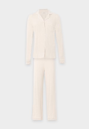 Femme aux longs cheveux foncés portant un haut beige clair à manches longues, boutonné, et un pantalon large assorti, debout pieds nus sur fond blanc.