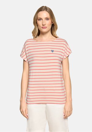 Blonde Frau trägt beige Kurzarmshirt mit Korallenstreifen und kleinem blauen Herz auf der Brust, kombiniert mit weißen Shorts, vor schlichter Hintergrund.