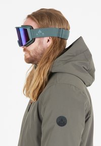 Hiihtolasit tummanvihreällä hihnalla, suuret peilipinnalla varustetut linssit purppuransävyisinä ja vaahtomuovinen vuori mukavuutta varten. Yhdistetty harmaaseen huppariin.