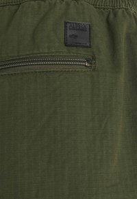 Olive zelená látka s diagonalným zipsovým vreckom a textúrovaným povrchom. Čierny štítok so šitým nápisom "ALPHA INDUSTRIES".