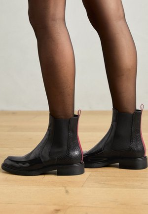 Beine in schwarzen, transparenten Strumpfhosen, die schwarze Leder-Stiefeletten mit elastischen Seitenteilen und pinkfarbenen Zugschlaufen tragen, stehen auf einem Holzboden.