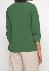 Green, tailored blazer with three-quarter sleeves, structured shoulders, and a straight fit, paired with light beige trousers.