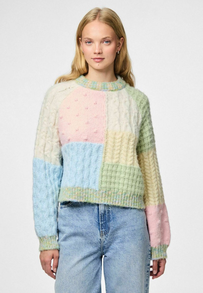 Kobieta nosząca sweter z kolorowego patchworku w pastelowych odcieniach, z wzorami warkoczy i fakturami, w zestawieniu z jasnoniebieskimi spodniami o wysokim stanie.