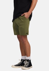 Pantalones cortos de color verde oliva con un ajuste relajado, cintura elástica y bolsillos laterales. Combinados con zapatillas altas negras y calcetines blancos hasta el tobillo.