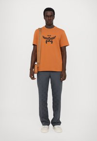 Oranžist puuvillane T-särk musta logoga, kokku sobitatud hallide mustriliste pükste ja kreemjate tossudega. Komplekta kuulub ka ristkeha kott pruuni rihmaga.