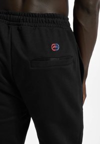 Black sweatpants in soft fabric with an elastic waistband. Features a small multicolored logo near the back pocket and a back pocket detail.
