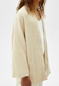 Person iklädd en lös, strukturerad beige kimono-liknande kappa över en vit topp, visad från bröstet och ner till handen mot en neutral bakgrund.