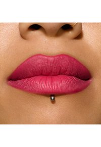 Nærbilde av fyldige lepper med matt rosa leppestift, jevn tekstur og en leppepiercing med en metallkule. Klar hud i bakgrunnen.