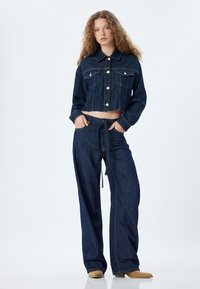 Chaqueta de denim con diseño cortado y bolsillos frontales, combinada con jeans de pierna ancha de tiro alto, ambos en un lavado oscuro, con un lazo en la cintura.