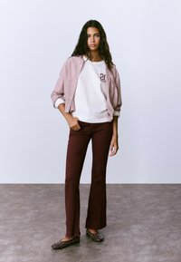 Mujer con cabello largo y oscuro, vistiendo una chaqueta de rayas rosa sobre una camisa blanca y pantalones burdeos, de pie sobre un suelo gris contra una pared blanca.