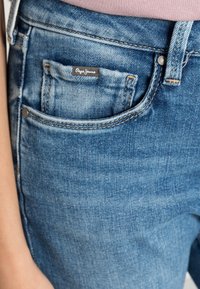 Jeans de ganga azul claro com um acabamento desbotado, apresentando um pequeno bolso frontal e o rótulo "Pepe Jeans". Detalhe de costura em contraste é visível.