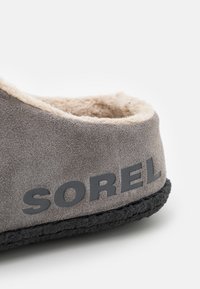 Sive pamučne papuče od sive antilop kože s bogatim kremastim podstavom, crnom gumenom potplatom i istaknutim logotipom "SOREL" sa strane.