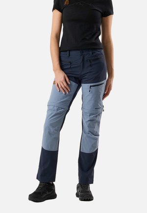 Kobieta ubrana w czarny t-shirt i dwukolorowe, niebieskie spodnie outdoorowe z kieszeniami na zamek, stojąca z ramionami swobodnie opuszczonymi wzdłuż ciała.