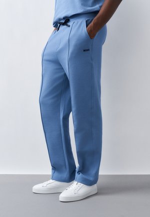 Mężczyzna w jasnoniebieskich spodniach dresowych z czarnym logo "BOSS" na udzie i białych trampkach, stojący z rękami w kieszeniach na jednolitym tle.