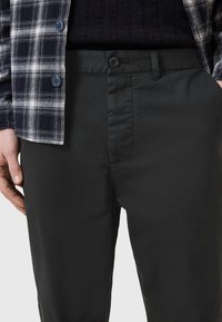 Muž v černých kalhotách se zapínáním na knoflík a bočními kapsami, v kombinaci s černým žebrovaným tričkem a černo-bílou kostkovanou koženou bundou.