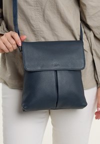 Ženska, oblečena v bež srajco in bele hlače, drži črno usnjeno torbico čez ramo z zaponko in vidnim logotipom.