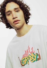 Camiseta blanca de algodón con un diseño gráfico colorido que presenta la palabra "GRIMY" sobre llamas estilizadas y figuras abstractas en marrón y verde.