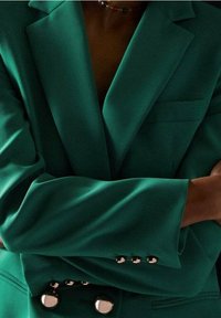 Teal-puku, joka on valmistettu sileästä kankaasta; siinä on leveät läpät, yksi rintatasku ja kiiltävät hopeanväriset napit hihansuissa ja edessä.