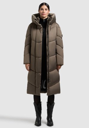 Dolga olivno zelena puhovka z visokim ovratnikom in kapuco. Ima prešite vzorce, zadrgo in stranske žepke. Nošena čez črno obleko.