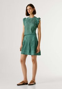 Frau steht und trägt ein ärmelloses grünes plissiertes Kleid und braune Sandalen vor einem schlichten hellen Hintergrund.