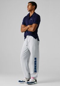 Man staat met gekruiste armen, draagt een marineblauw poloshirt, lichtgrijze joggingbroek met de tekst "REBEL", en meerkleurige sneakers op een grijze achtergrond.