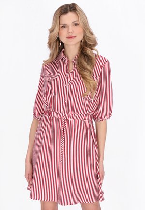 Donna bionda che indossa un vestito abbottonato a righe verticali rosse e bianche, con vita stretta e maniche fino al gomito, in piedi contro uno sfondo bianco.