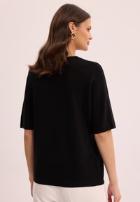 Czarny sweter z krótkim rękawem wykonany z dzianiny, o luźnym kroju i teksturowanej powierzchni, zestawiony z jasnymi spodniami. Prosty, czysty design.