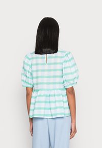 Top de mangas bufantes em azul claro com padrão xadrez verde e branco. Apresenta um acabamento peplum e um detalhe de abertura na parte de trás, tecido texturizado.
