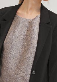 Žena v texturovaném béžovém topu pod černým sakem s jedním knoflíkem, odhalující svůj krk a částečnou čelist.