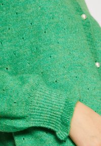 Zelený pletený cardigan s texturovanou tkaninou, s malými otvory, žebrovanými manžetami a knoflíky podobnými perlám na přední straně.