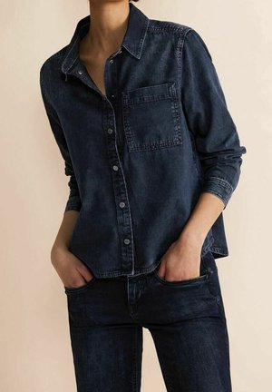 Personne portant une chemise en jean foncé à boutons avec poche poitrine et jean foncé assorti, mains dans les poches, sur fond uni.
